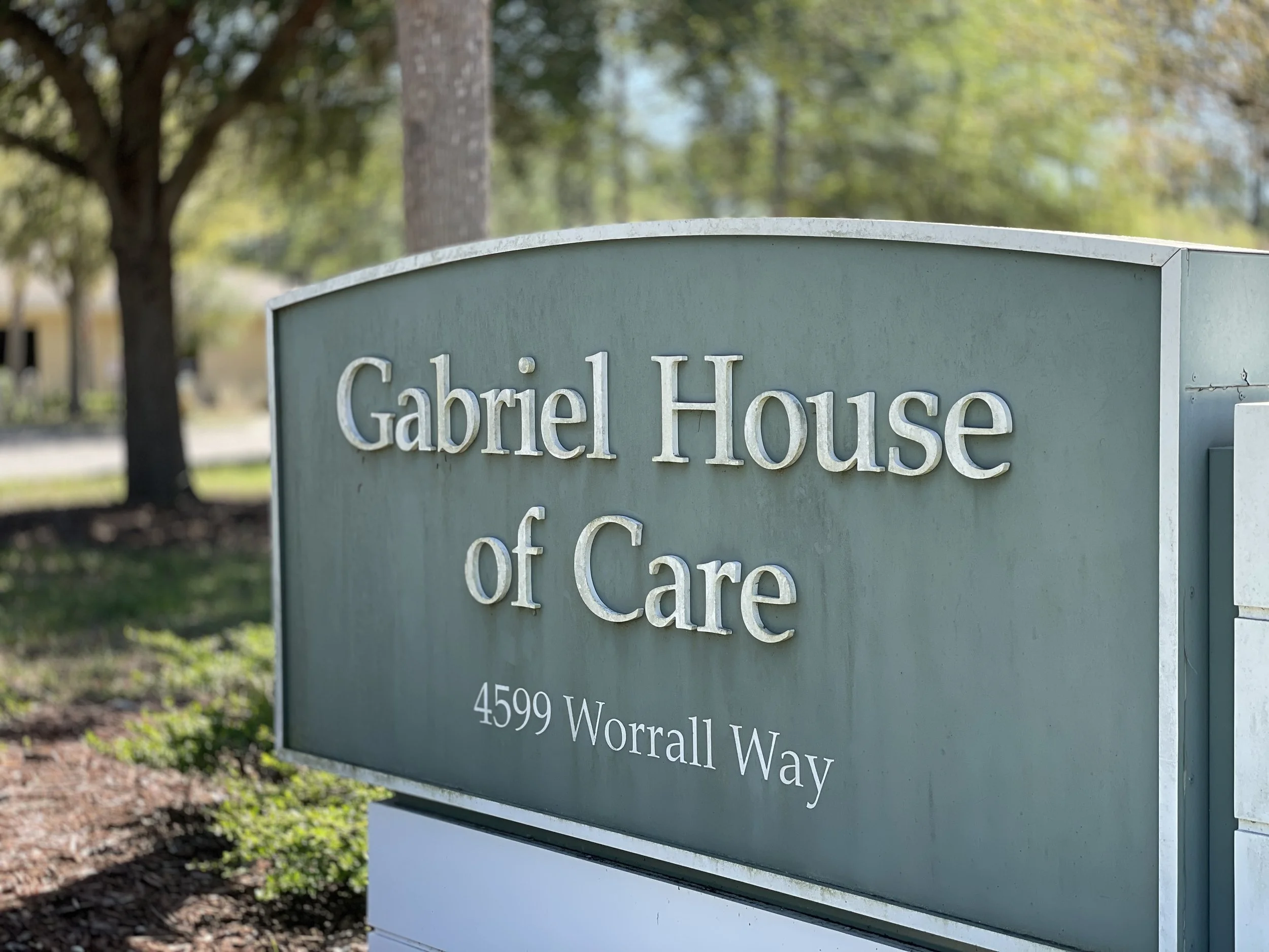 Street sign reading "Gabriel House of Care" with an address "4599 Worall Way" in a green background. Trees and a building are visible in the background.