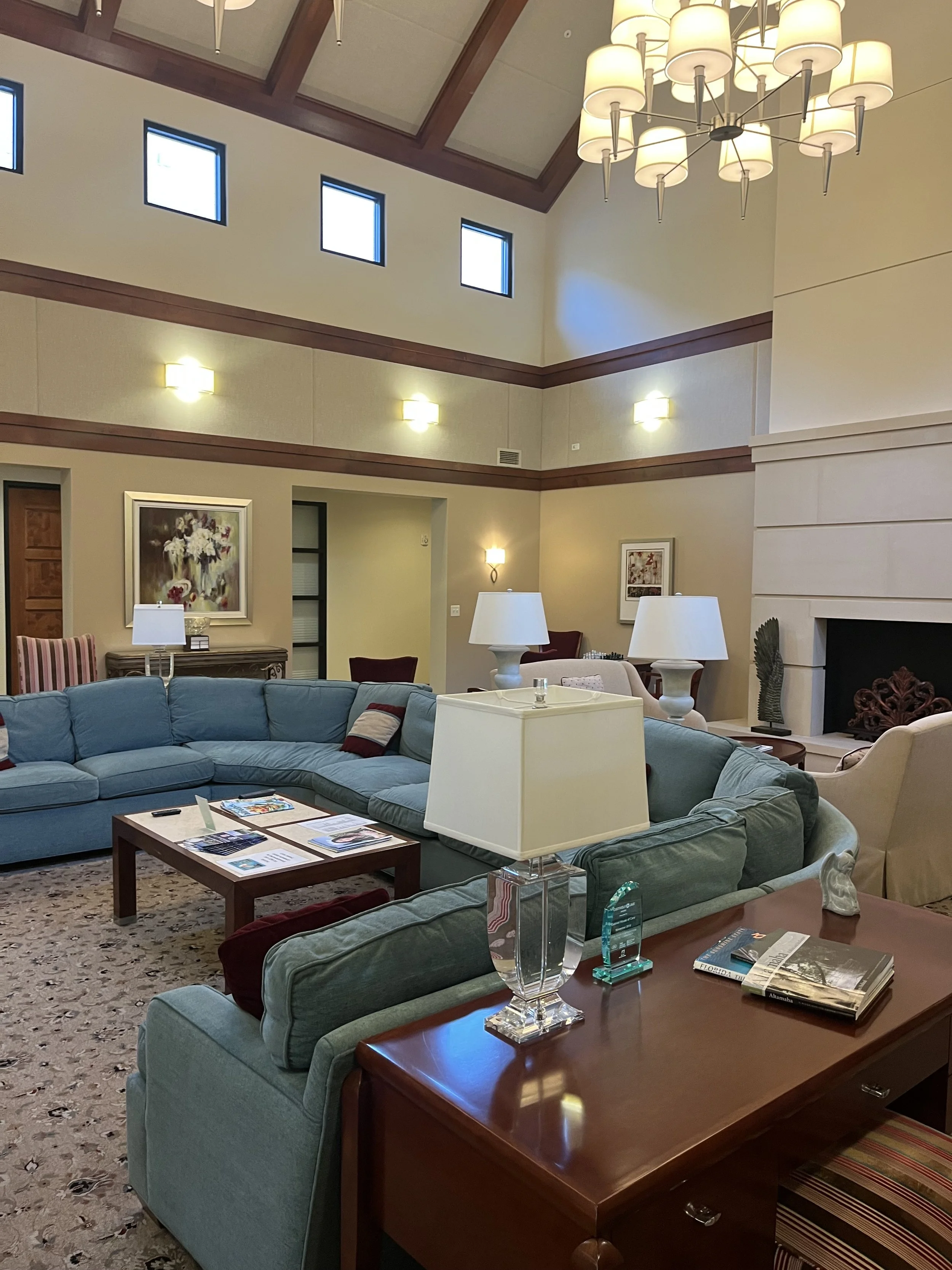 Living room with a blue sectional sofa, wooden coffee table with magazines, cream-colored walls, artwork, lamps, and a large chandelier hanging from the ceiling.