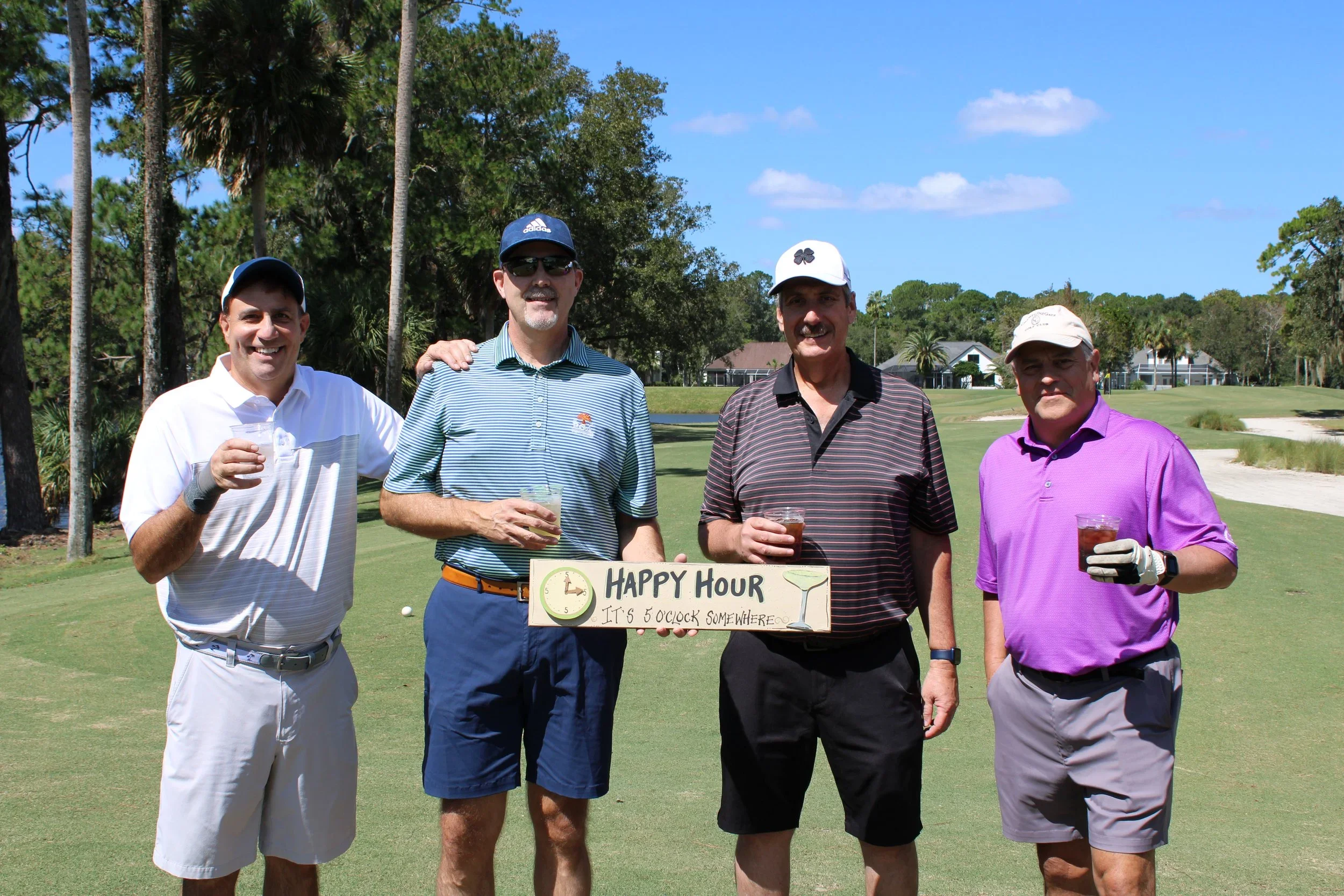 Four golfers smiling and raising a toast by a "Happy Hour" sign on the course during Gabriel House’s annual golf tournament.