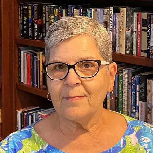 An older woman with short gray hair, black glasses, and hoop earrings, wearing a colorful floral top, standing in front of a bookshelf filled with books.