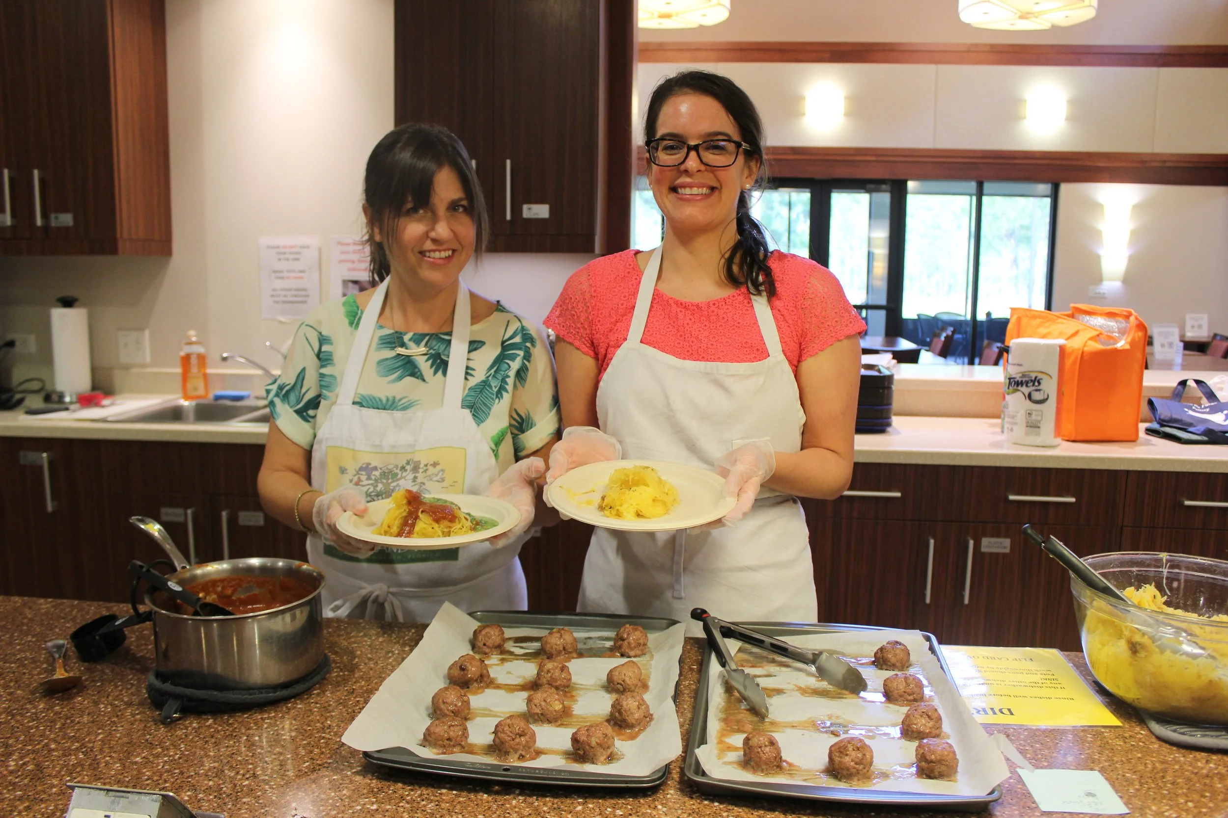 Two women standing behind a kitchen counter, holding plates of food, with a tray of meatballs and a saucepan in front of them. They are smiling and wearing aprons, with a kitchen and dining area in the background.