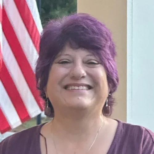 A woman with short purple hair smiling outdoors, with an American flag in the background.