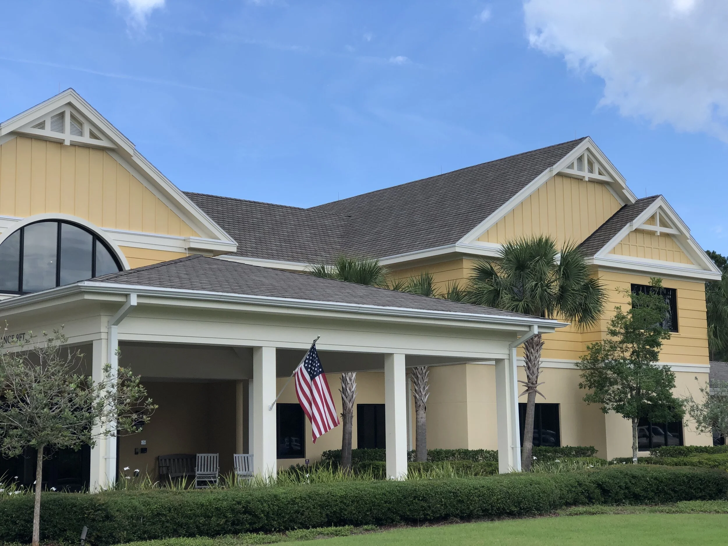 A yellow house with white trim and a dark gray roof, surrounded by palm trees, with an American flag hanging on the front porch, green grass, shrubs, and a blue sky with some clouds.