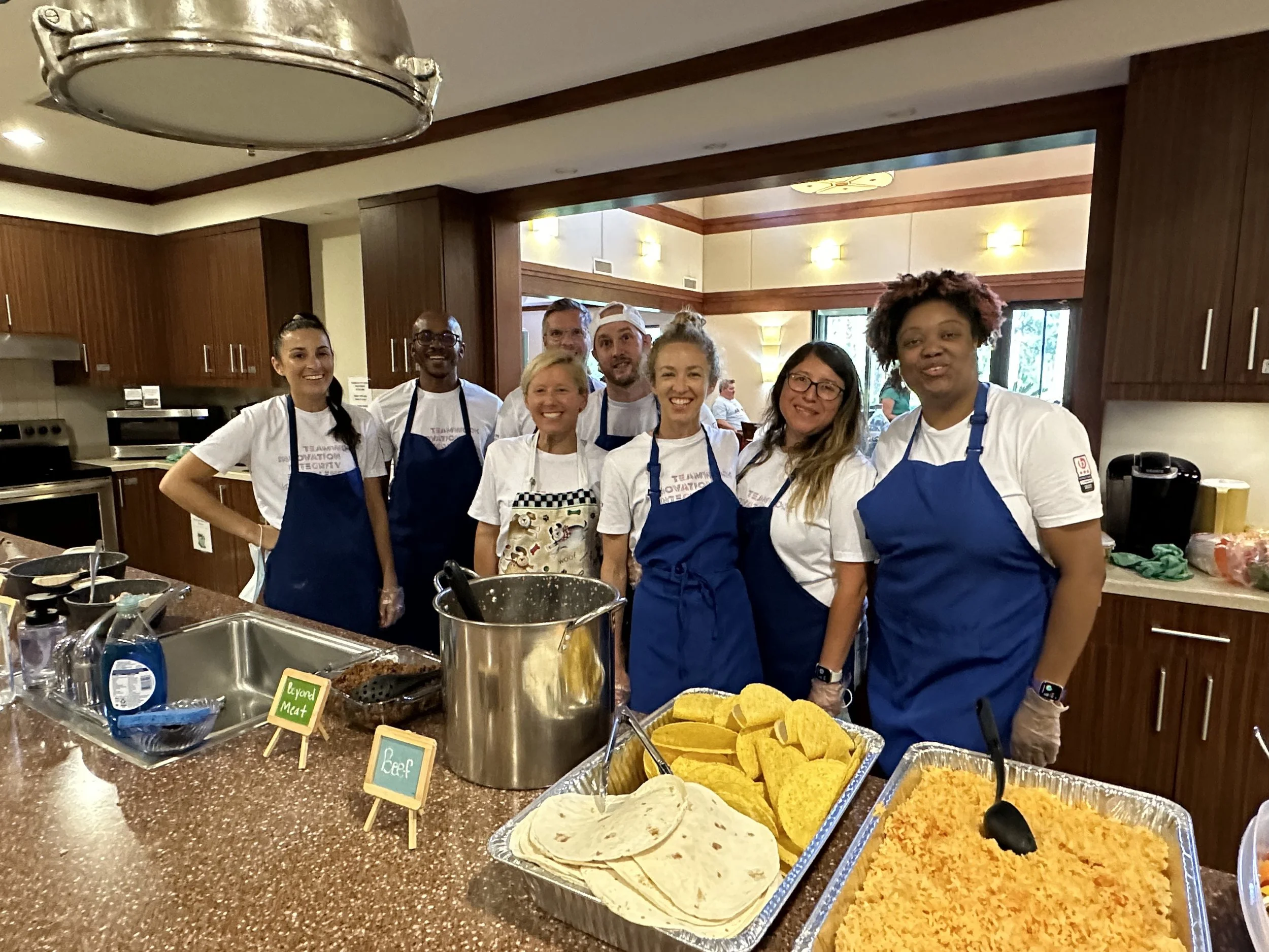 A group of diverse people wearing chef uniforms and aprons standing together in a kitchen, smiling for a photo.