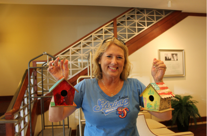 A woman holding two colorful birdhouses, smiling indoors with a staircase and framed picture on the wall in the background.