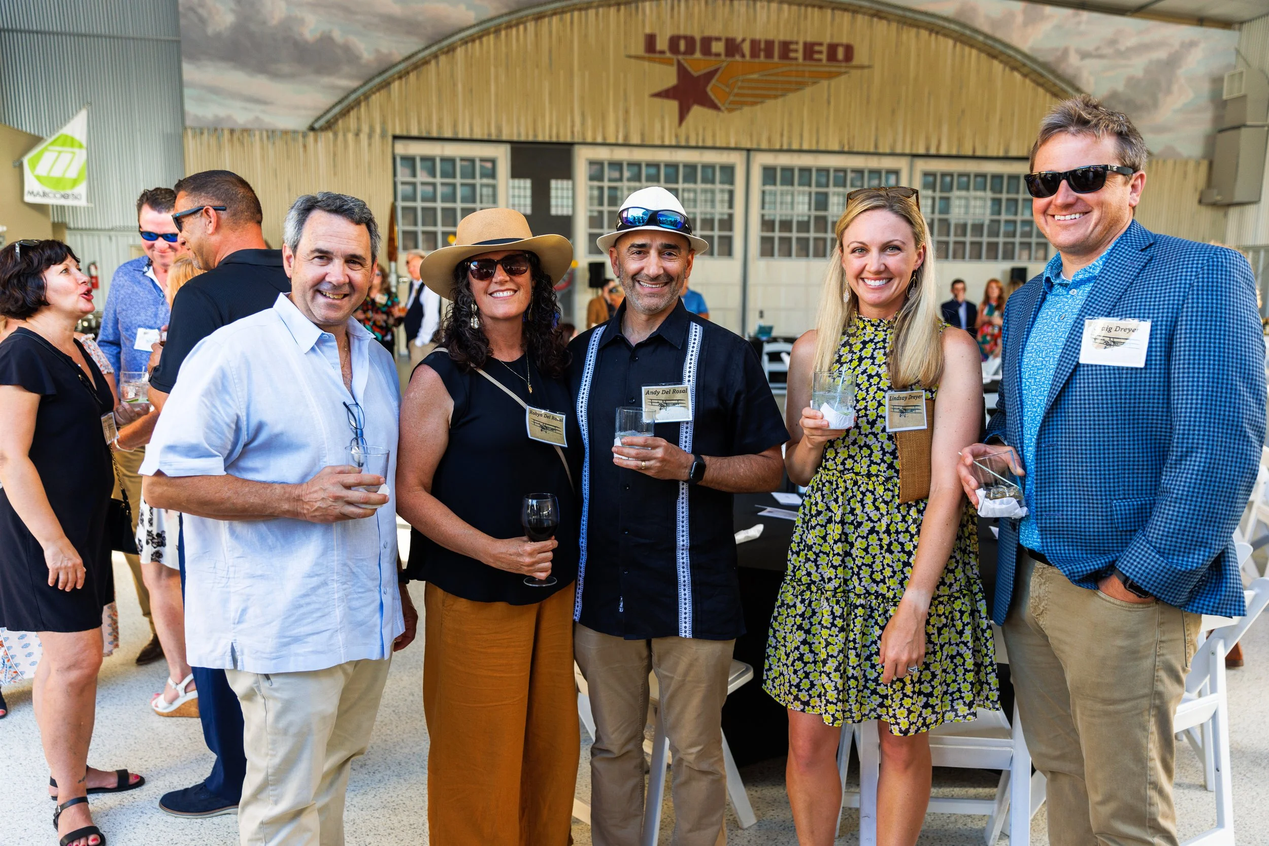 Group of six smiling adults standing together at an event in front of a building with the Lockheed logo, holding glasses of wine, with others mingling in the background.