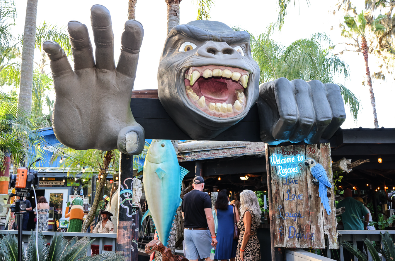 Large decorative sculpture of a gorilla with an open mouth, showing teeth and fangs, holding a hand up with fingers spread wide. The scene is outdoors with palm trees and people around, appearing to be at a lively restaurant or bar entrance.
