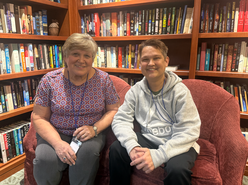 Woman and young man sitting on a red couch in front of a bookshelf filled with books, both smiling at the camera.