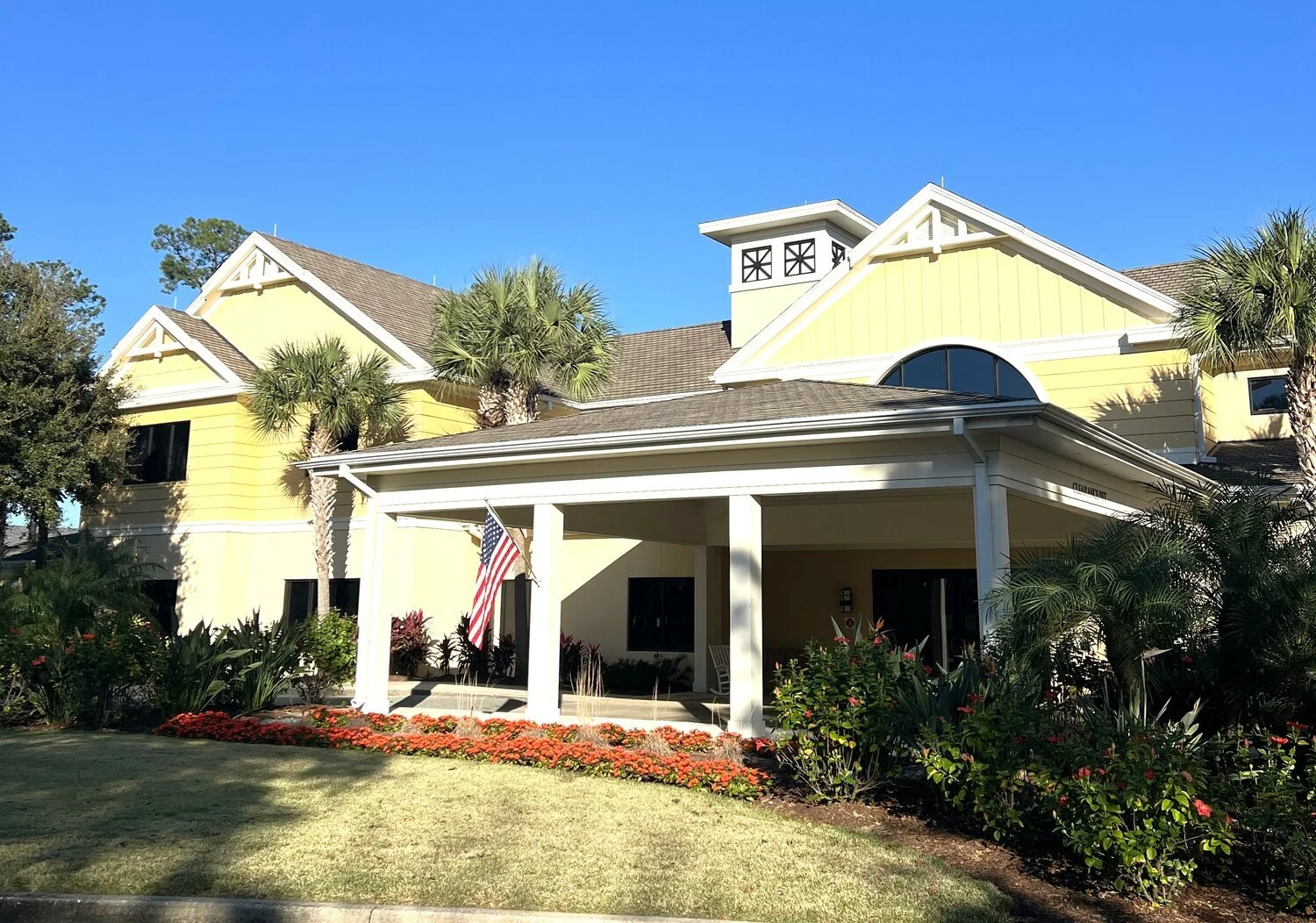 A yellow house with white trim and a brown shingle roof, surrounded by palm trees, with an American flag hanging outside. The front yard has green grass and red flowers, under a clear blue sky.