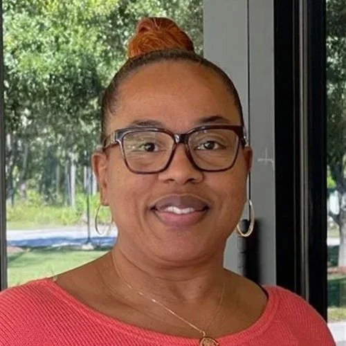 A woman with glasses, hoop earrings, and a bun hairstyle standing indoors near a window with trees outside.