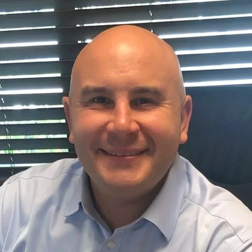 A smiling bald man in a light blue button-up shirt, sitting in front of window blinds.