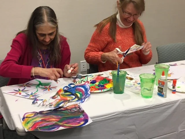 Two women sitting at a table engaged in arts and crafts, with colorful paints, scissors, and glue on the table, smiling and cutting paper.