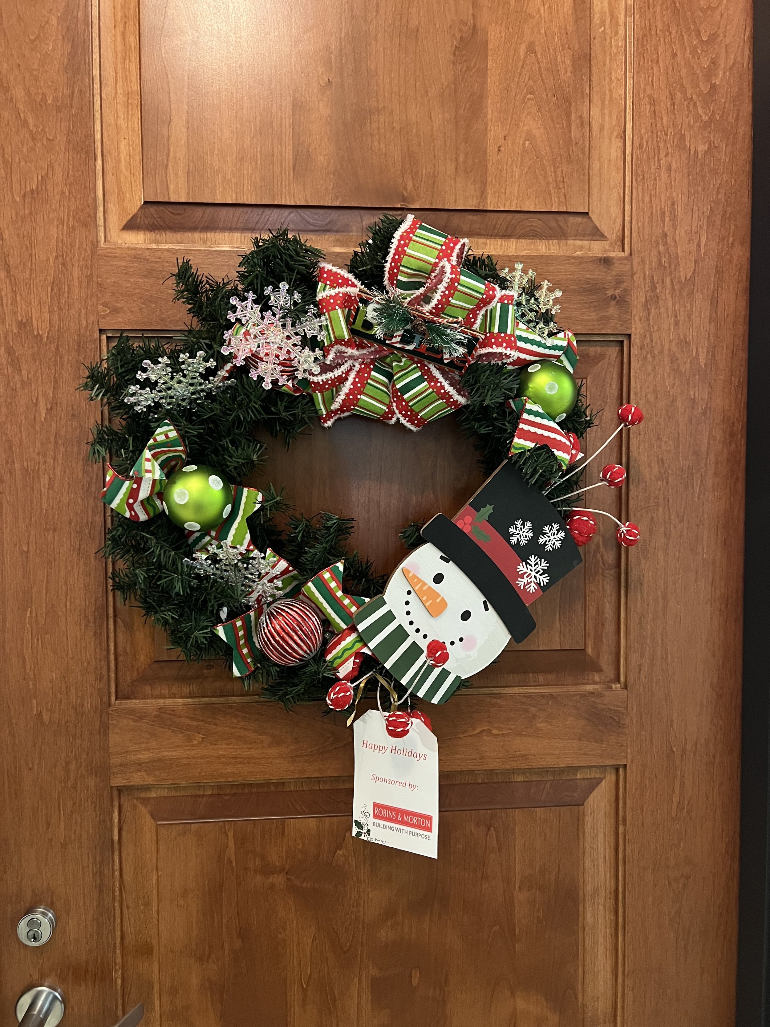 A Christmas wreath decorated with striped ribbons, shiny green ornaments, snowflake ornaments, and red berries, hanging on a wooden door with a white holiday tag attached.