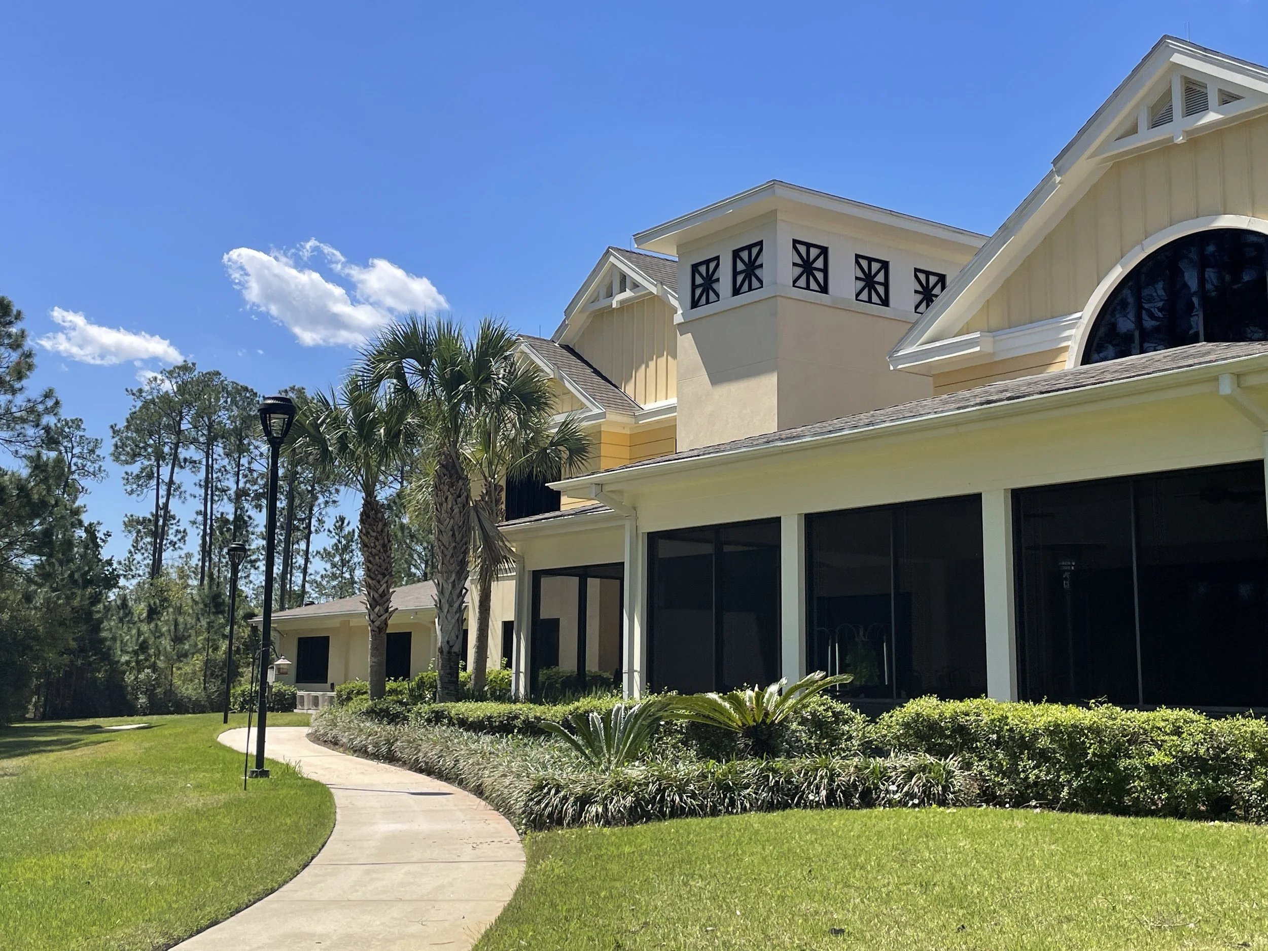 A yellow residential house with large windows, surrounded by neatly trimmed greenery, palm trees, and a sidewalk, under a bright blue sky with scattered white clouds.