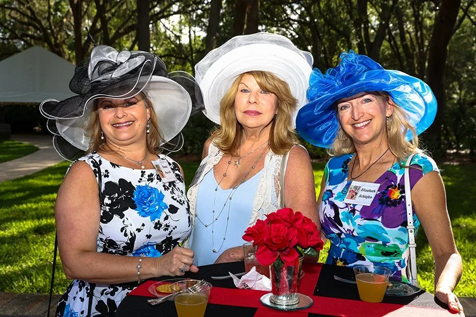 Three women wearing large, elaborate hats and floral dresses standing outside at a table with red flowers in a glass vase, drinks, and a dessert, smiling.