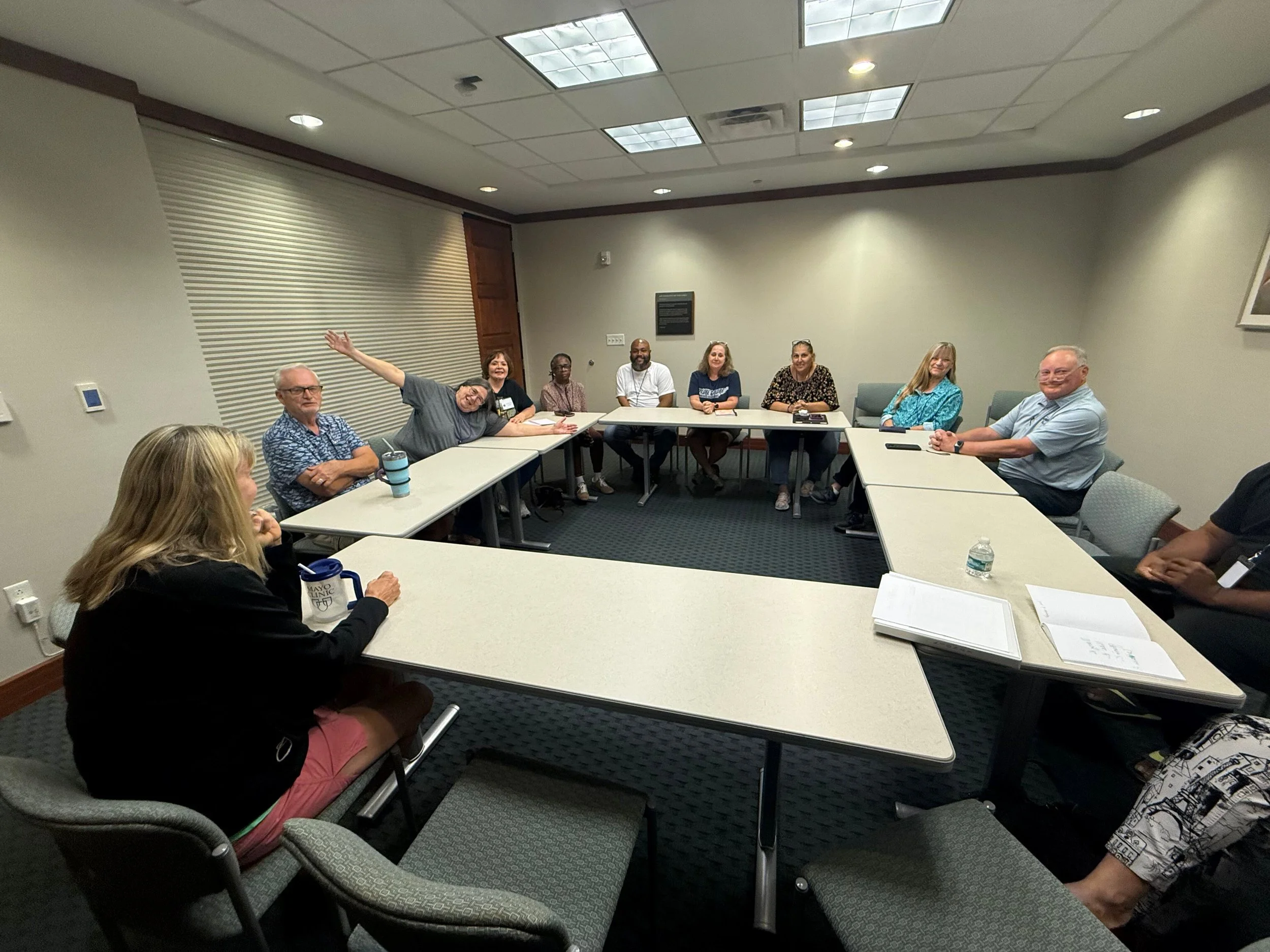 A group of ten diverse people sitting around a large U-shaped conference table in a well-lit meeting room, smiling and engaging in a discussion.
