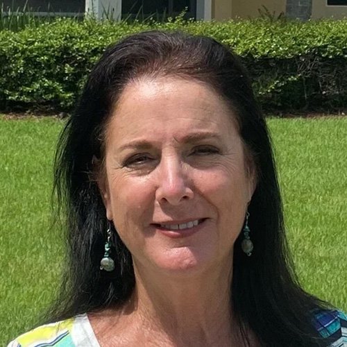 A woman with long dark hair wearing earrings, standing outdoors on a sunny day with green grass and bushes in the background.