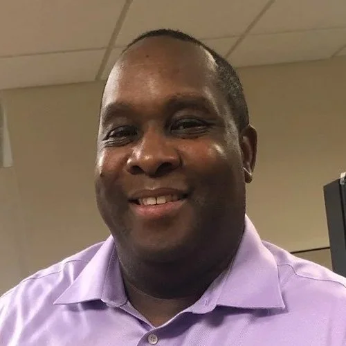 A smiling man wearing a light purple dress shirt indoors with a ceiling and a computer monitor in the background.