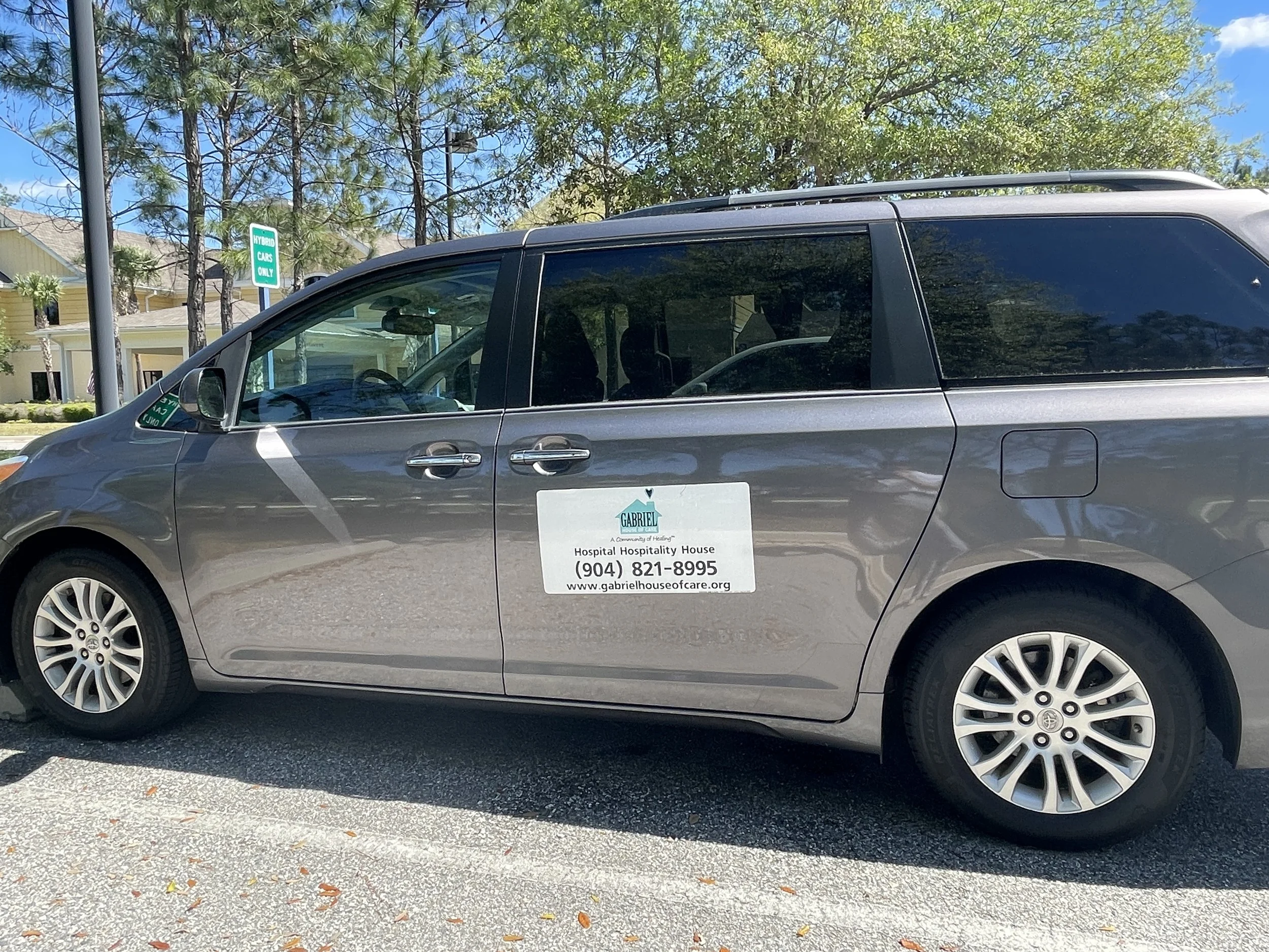Silver hybrid car parked outdoors with a sign on the side reading 'Gabriel Hospital Hospitality House' including a phone number and website. Background shows trees, blue sky, and residential buildings.