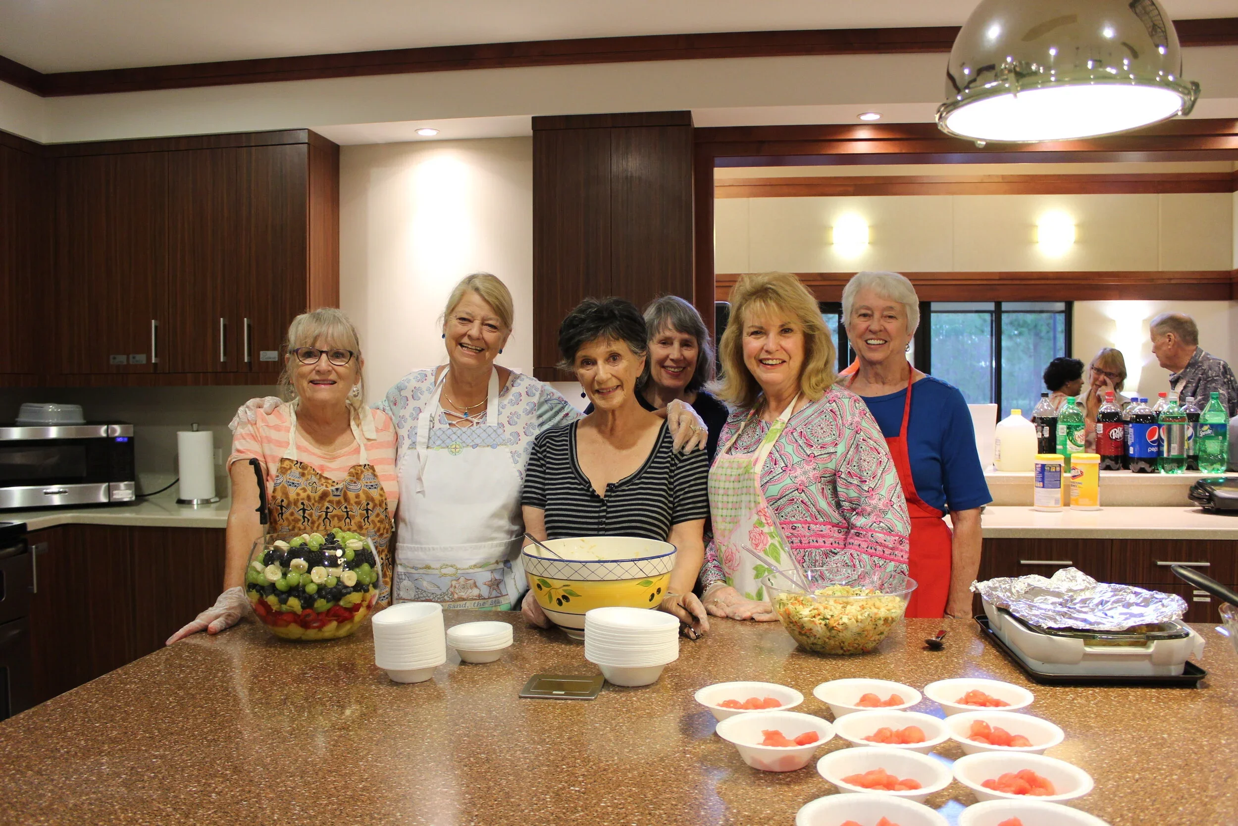 Group of smiling women in a kitchen preparing food and drinks for a gathering, with bowls of salad and fruit, and bottles of soda on the counter.