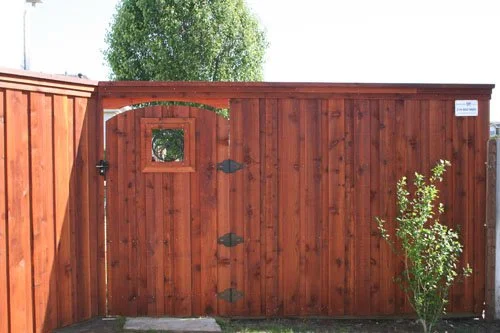 Wooden backyard gate with a small square window and decorative metal hinges, next to a shrub.