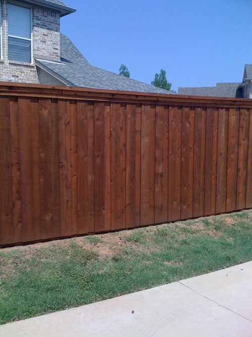 A wooden privacy fence with vertical planks in front of a house with brick siding and a pitched roof. The sky is clear and blue.
