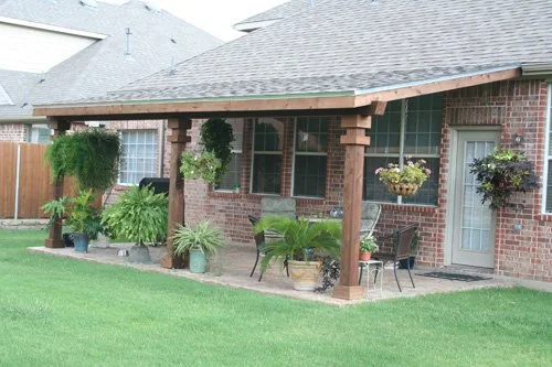 Backyard patio with potted plants, hanging baskets, patio table, and chairs under a roof extension of a brick house.