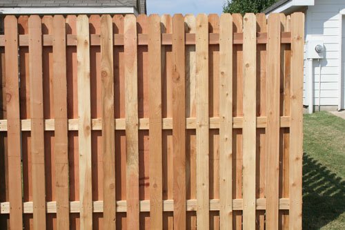 A wooden privacy fence with vertical planks and horizontal supports, installed in a backyard with green grass and a corner of a white house visible.