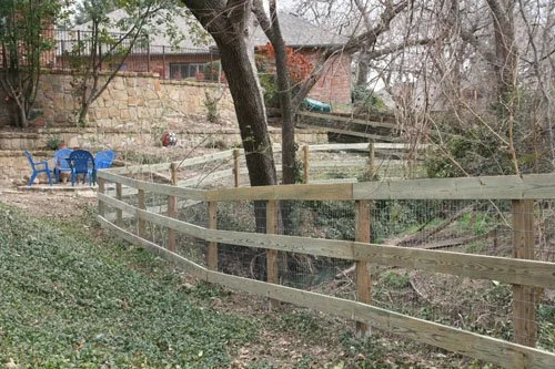 A backyard with a wooden fence, a tree, a stone wall, and a few blue chairs on a grassy area.