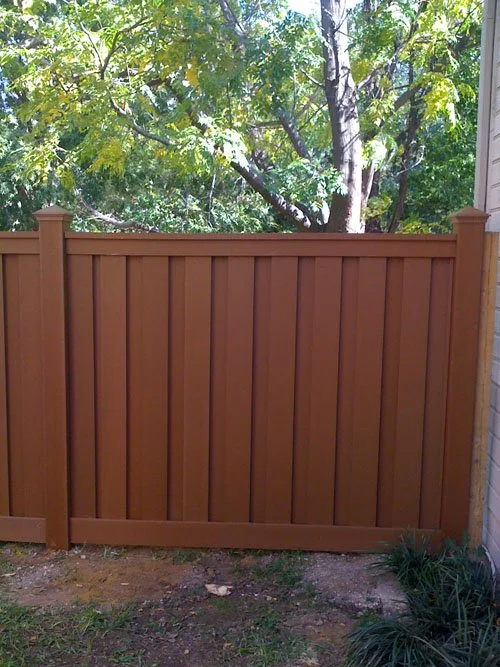 A wooden privacy fence with vertical planks and decorative posts, with trees and greenery in the background.