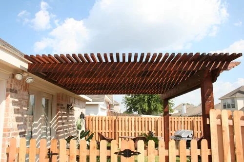 Wooden patio cover over backyard seating area with a brick house, fence, and trees in the background.