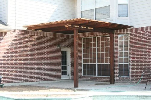Backyard patio with a wooden pergola attached to a brick house, showing a door and windows.