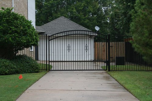 A black metal gate in front of a driveway leading to a garage with a white door, surrounded by green grass, bushes, and trees.