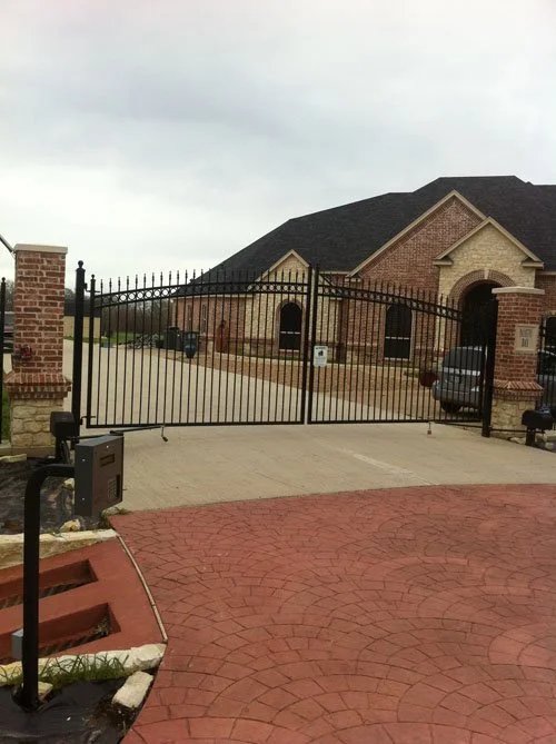 A black metal gate blocking a driveway in front of a brick house with a gable roof, brick columns, and a concrete driveway, in a residential neighborhood.