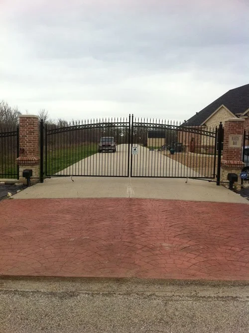 View of a gated driveway with black metal gates, brick pillars on each side, concrete and brick paving, and a house in the background.