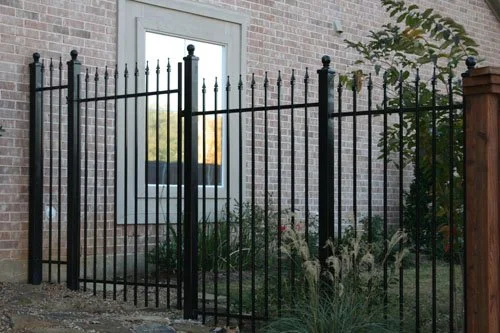 A black metal fence in front of a brick house with a window, some greenery, and a bush.