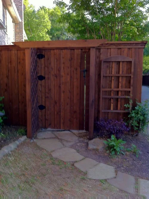 A wooden backyard gate with a curved top on a stone pathway, surrounded by plants and trees.