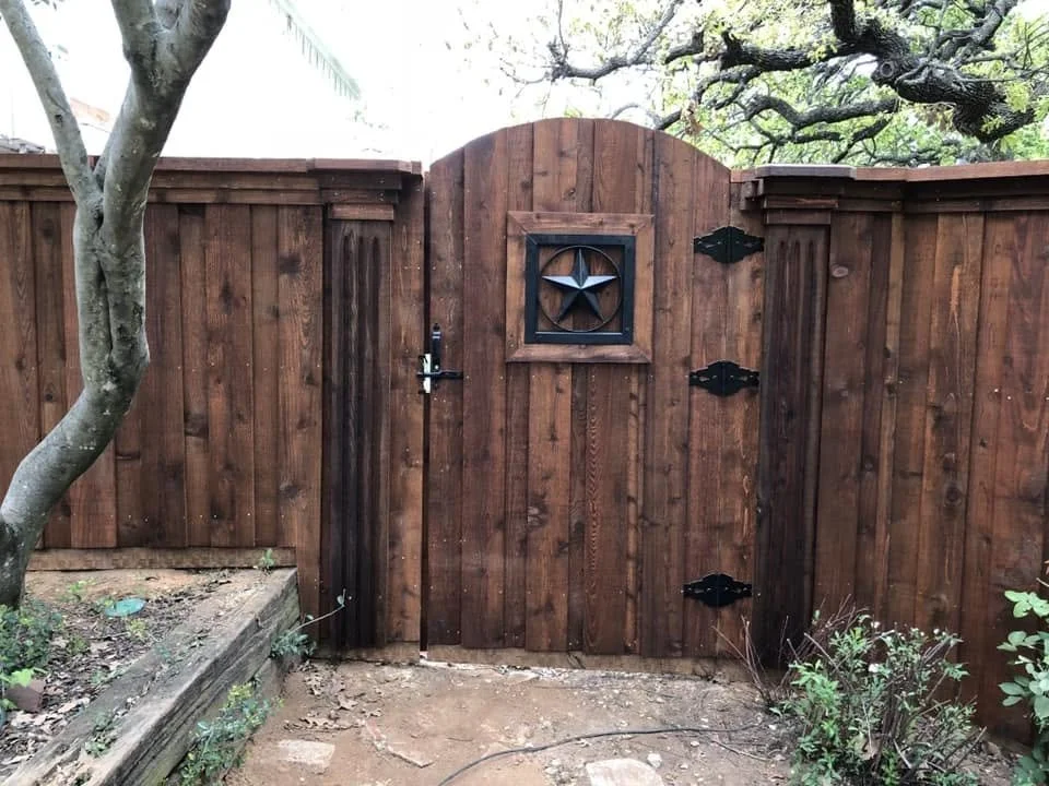 A wooden backyard fence with a small door, a star-shaped decoration, and a fish-shaped metal art piece. There are trees and plants nearby.