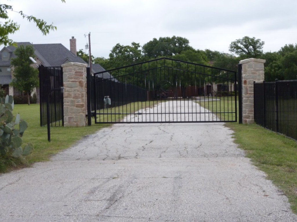 A black metal gate with stone pillars on either side, closed across a gravel driveway leading into a yard with grass and trees, with a house visible in the background.