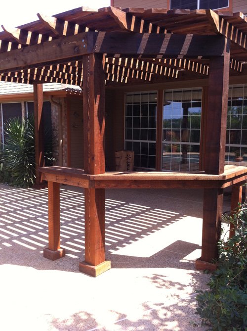 Wooden outdoor pergola with slatted roof and railing, casting striped shadows on the ground in front of a house with large windows and greenery.
