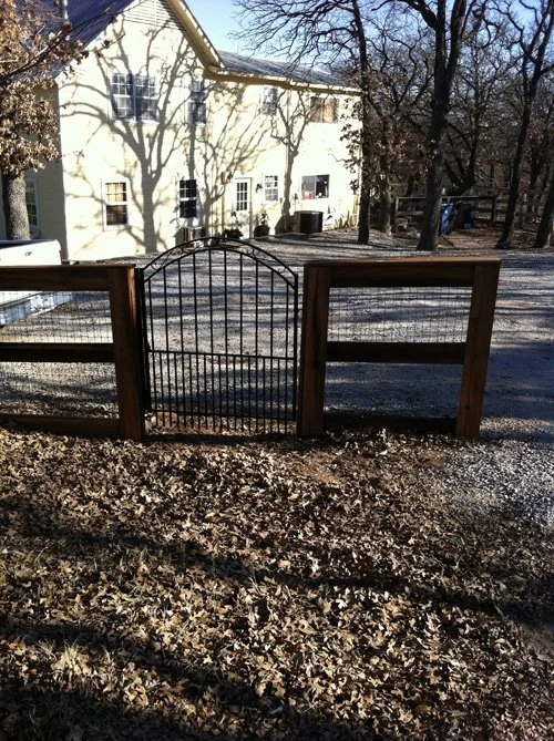 A small black metal gate with a curved top, set between two wooden fence posts, in front of a house with a light yellow exterior and multiple windows, surrounded by leafless trees and a dirt yard.