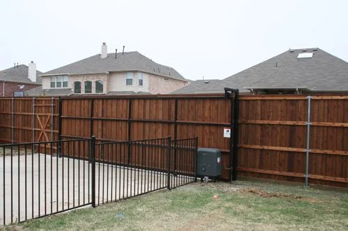 Backyard with a wooden fence, metal gates, and a small grass area, with neighboring houses in the background.
