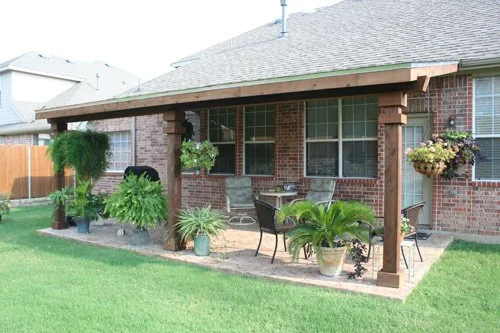 Backyard patio with seating area, potted plants, hanging flower basket, and a brick house with windows.
