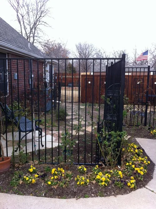 Backyard with a black metal gate, patio chairs, a stone walkway, flower beds with yellow and purple flowers, a wooden fence, and an American flag.