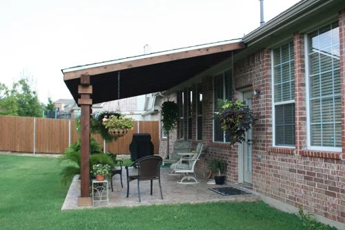 Backyard patio with outdoor furniture, hanging flower basket, and potted plants, next to a brick house with large windows.