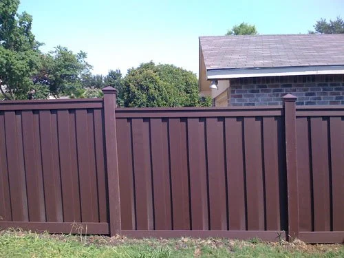 Brown wooden fence in front of a house with a sloped roof and trees in the background.