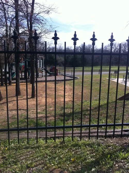 A fenced playground area with trees, a slide, and play structures, set in a park with grass and walking paths under a cloudy sky.