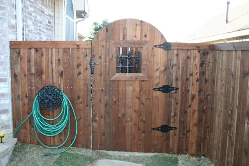 A wooden backyard fence with a small arched gate and decorative black hinges, with a garden hose hanging on the left side.
