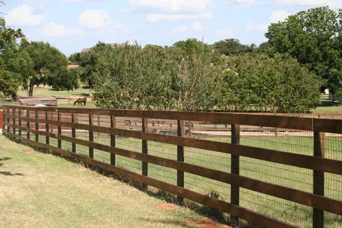 A wooden farm fence with horizontal planks and wire mesh, extending along a grassy area with trees and horses in the background under a partly cloudy sky.