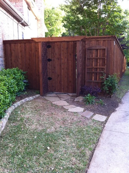 A wooden backyard privacy fence with a gate, surrounded by grass, bushes, small plants, and trees, with a sidewalk on the right side.